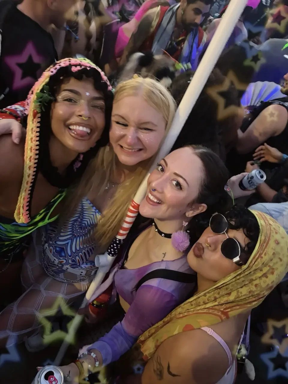 Four friends with face gems, smiling together in the crowd at a festival